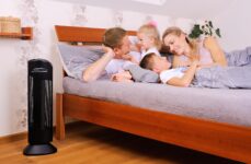 Čistička vzduchu Ionic-CARE v spálni vedľa postele, v ktorej sa rozprávajú matka, otec a ich dve malé deti, farba čierna